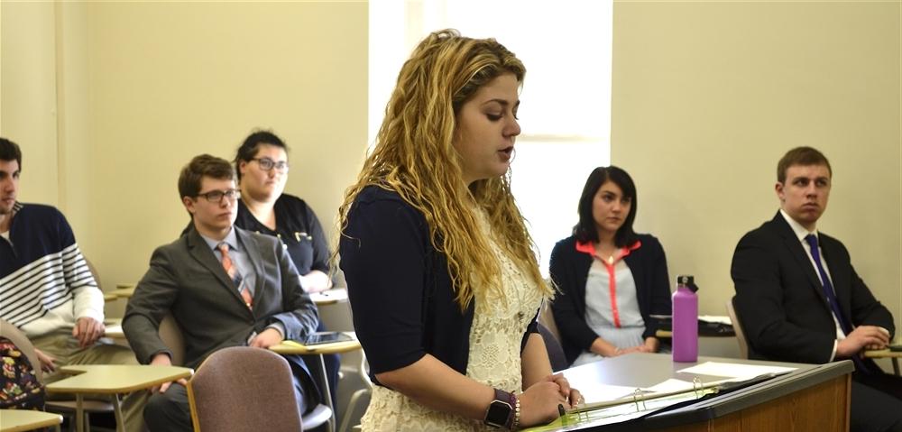 Students in a classroom and one student standing at a podium talking 